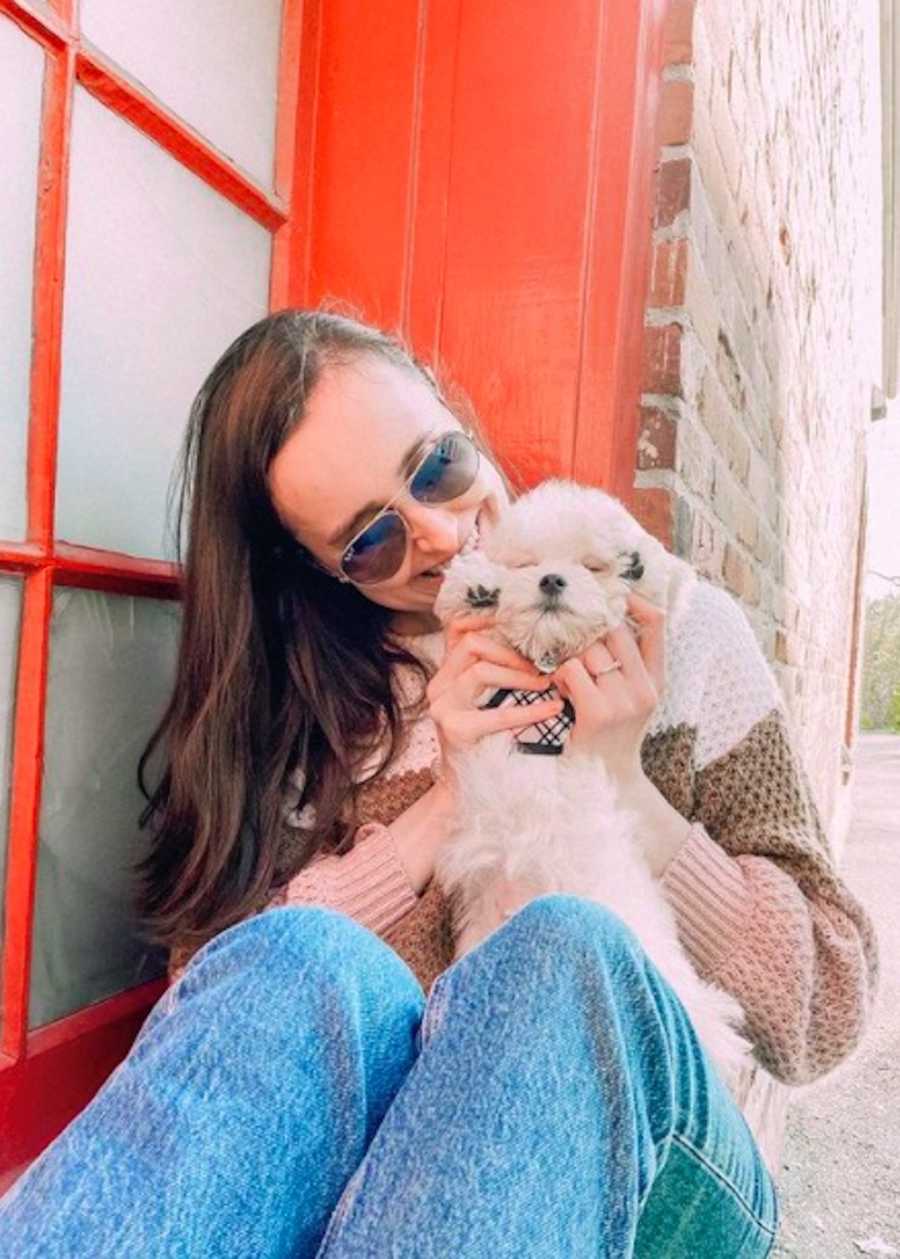 Young woman wearing sweater and sunglasses holding small white dog