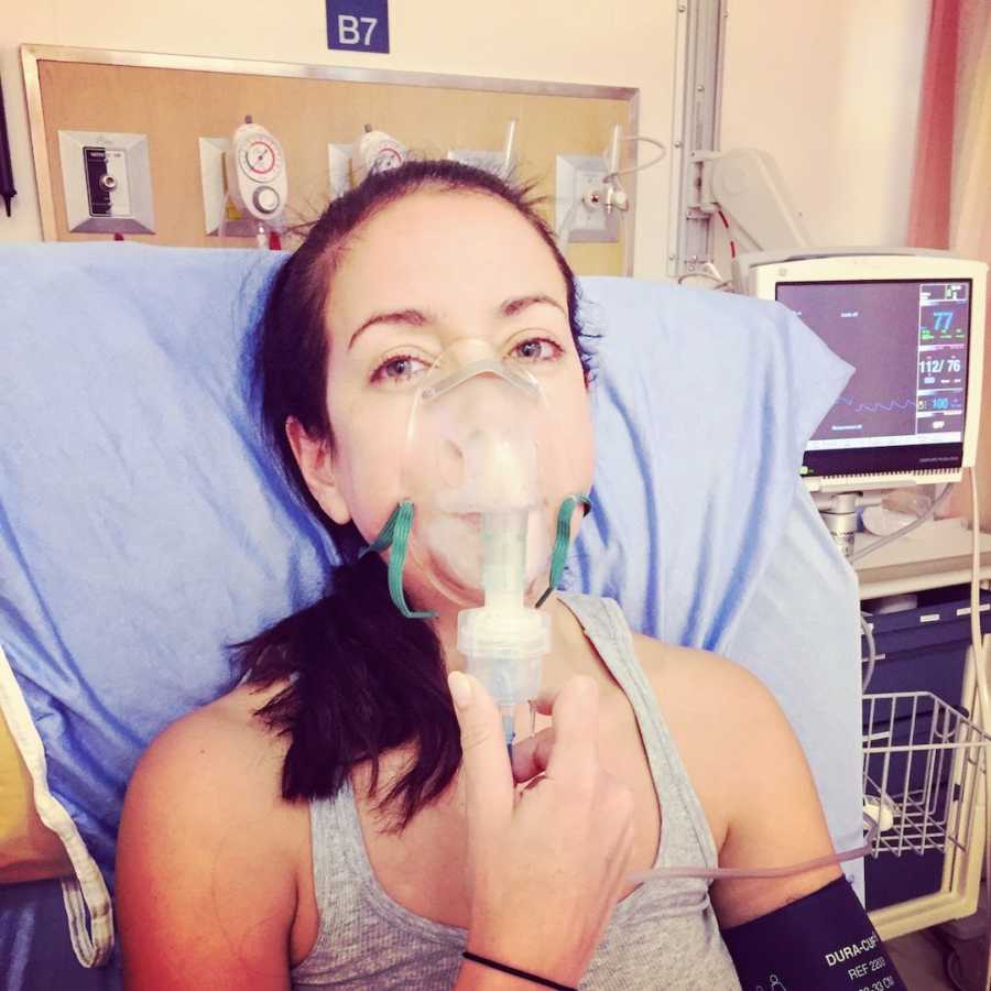 Woman wearing gray tank top with oxygen mask on face in hospital bed