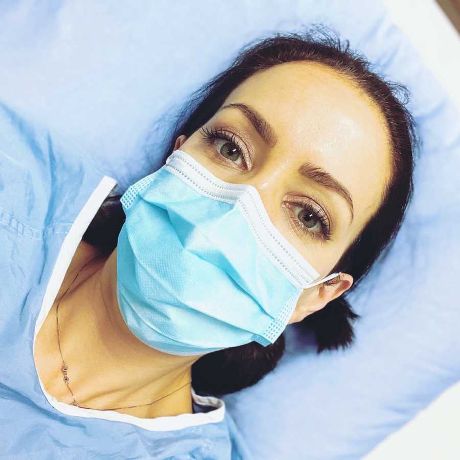 Woman lying down in hospital bed wearing hospital gown and face mask