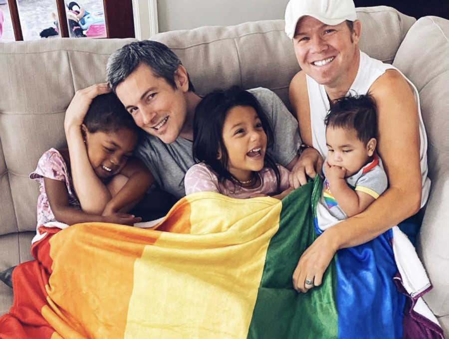 Two gay dads holding their three mixed race, adopted children and a rainbow flag