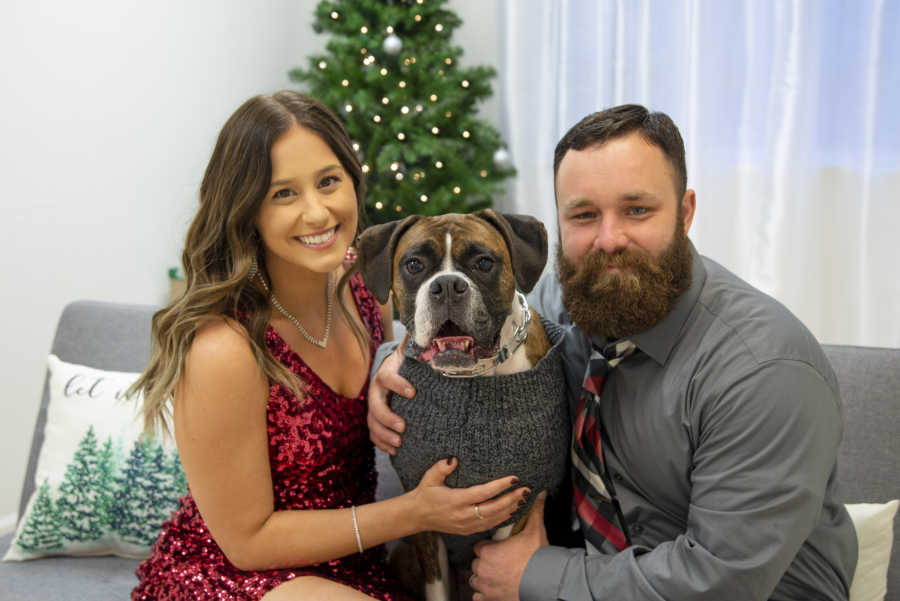 Husband and wife in front of Christmas tree with Boxer dog in sweater