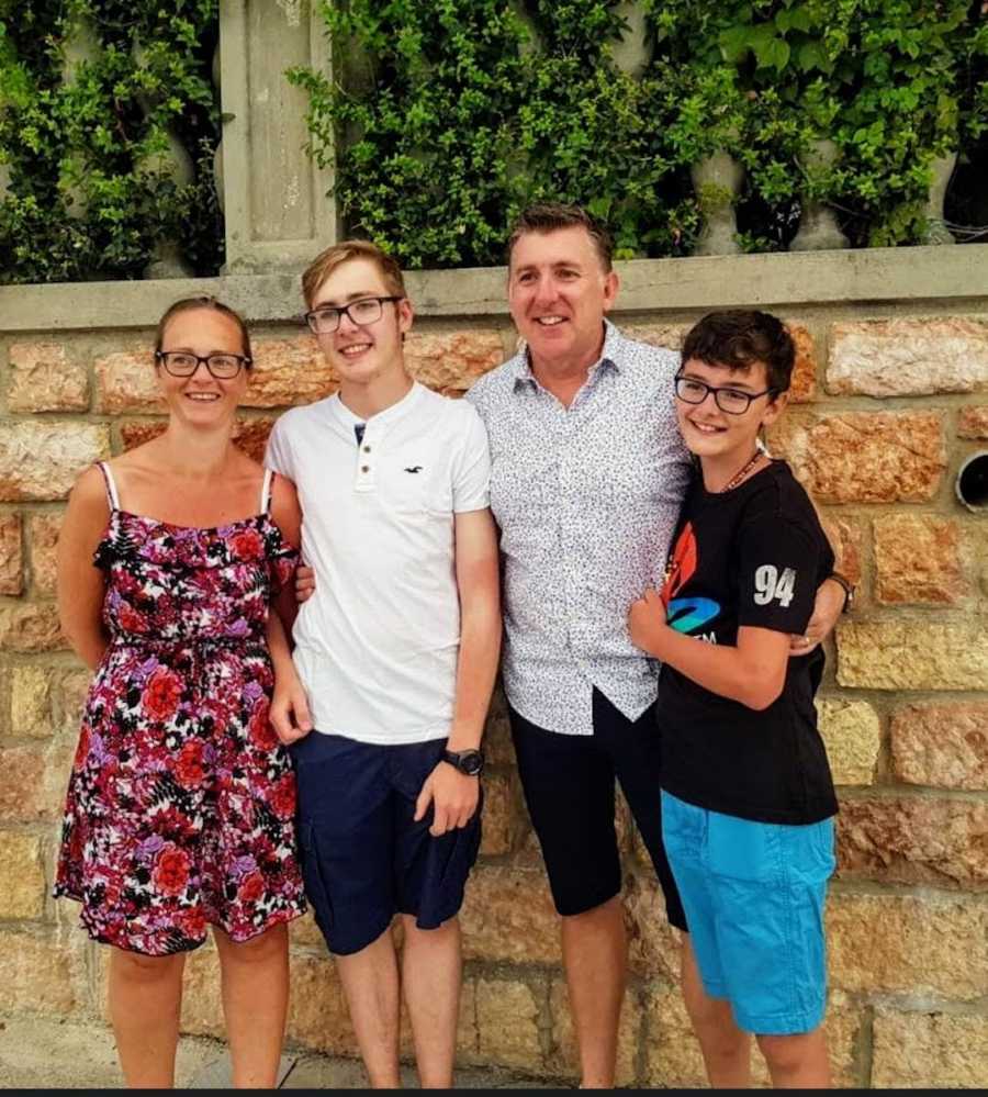 Family of four with arms around each other smiling in front of stone wall