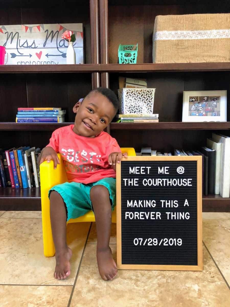 Young boy sits in yellow chair holding a letterboard with an adoption announcement
