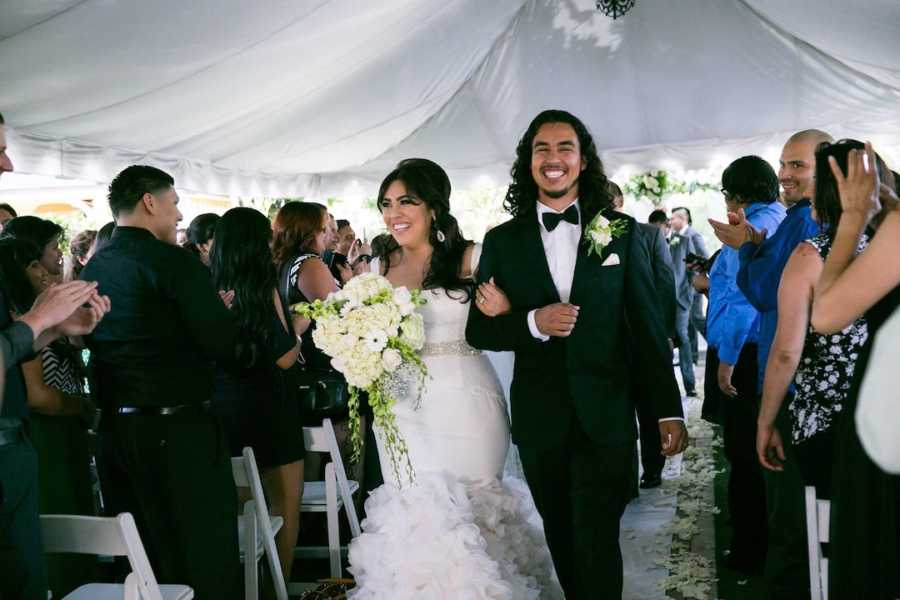 Newlywed couple walking down aisle holding flowers with cheering guests