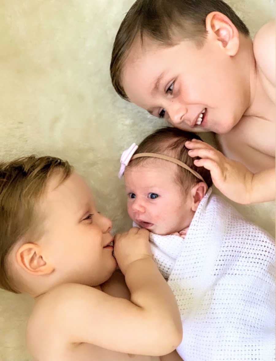 Two brothers and newborn sister wrapped in white blanket lying on floor and smiling at each other