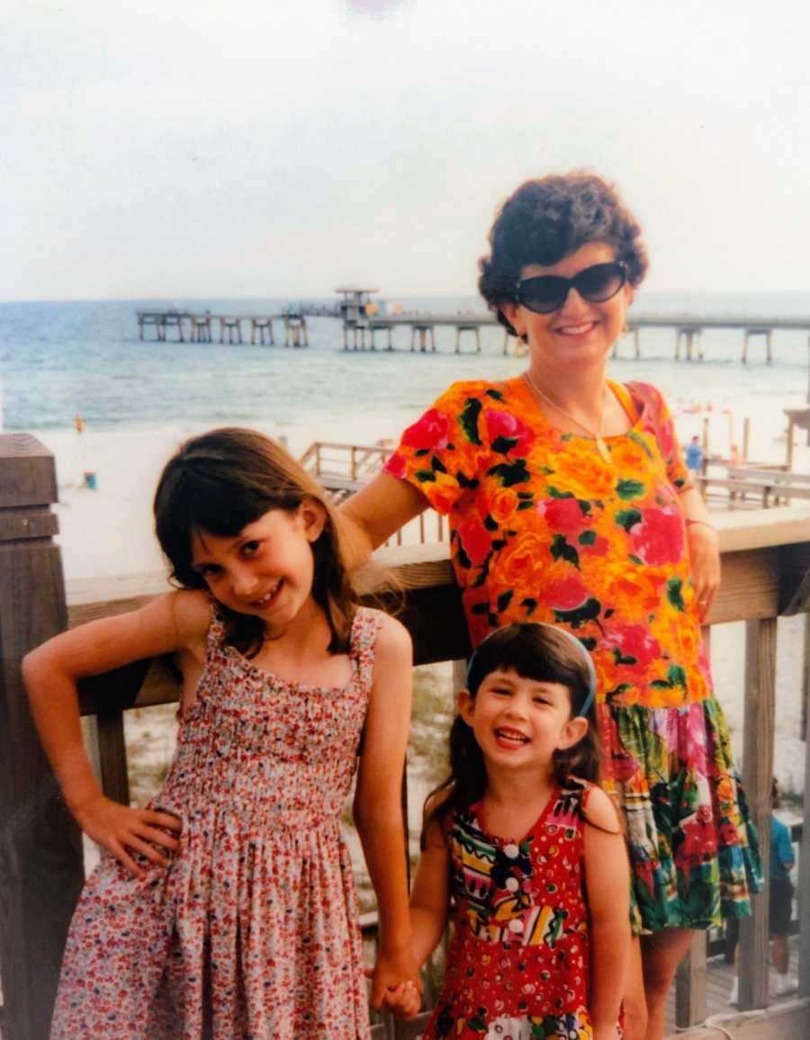 A mother stands in front of a beach pier with her two young daughters