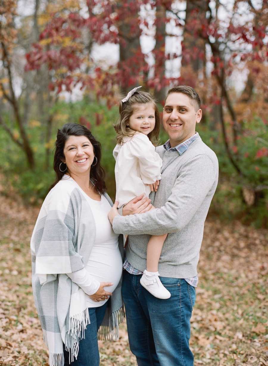 A family of three with pregnant mother stand in front of a woods smiling