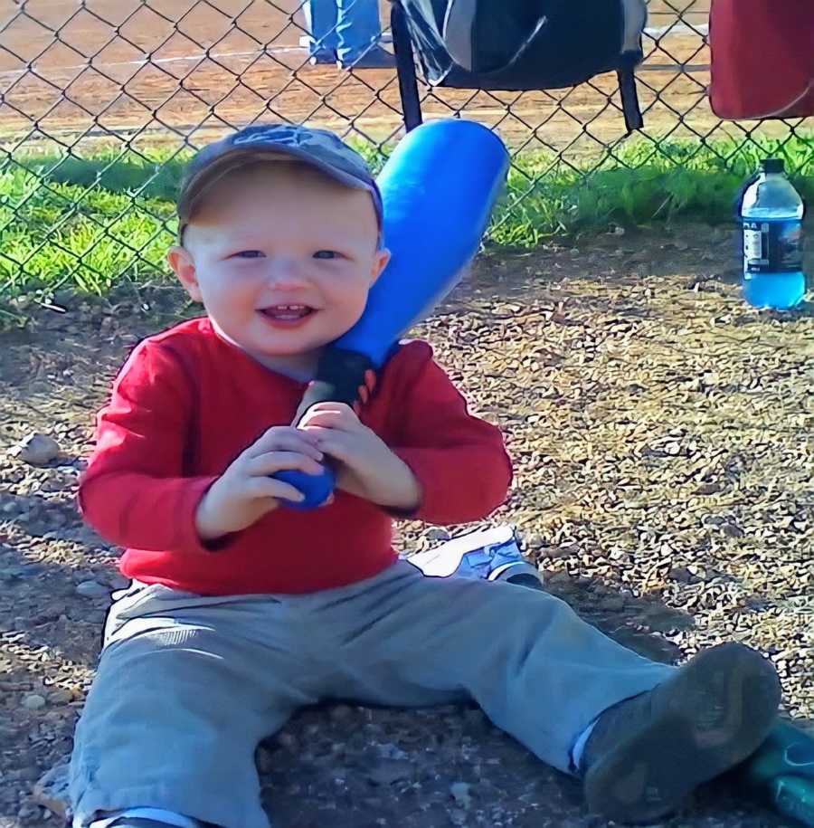 A boy wearing a baseball cap sits in the dirt holding blue baseball bat