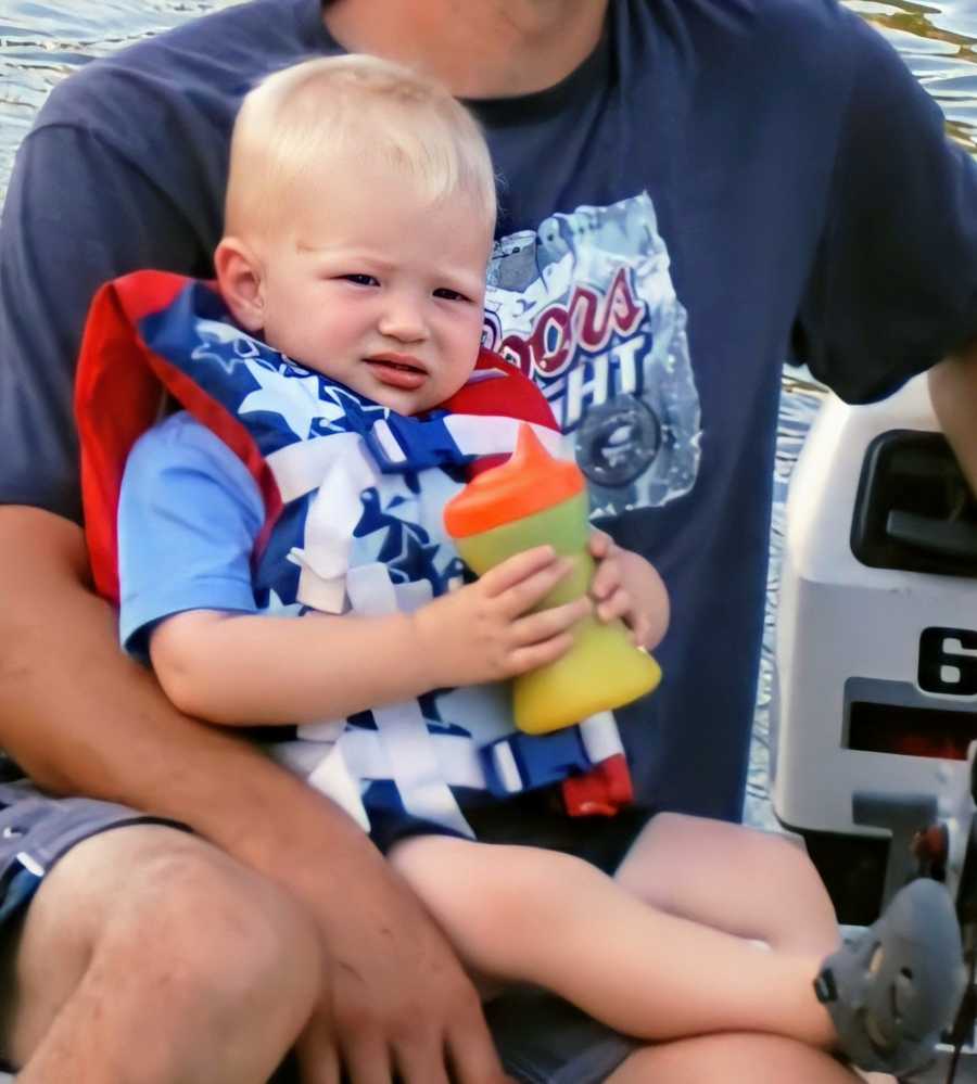 A boy wearing a life vest holds a sippy cup while on a man's lap