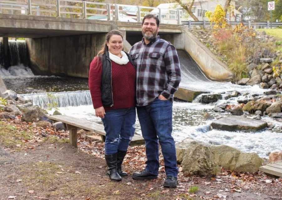 A married couple stand together outside by a waterfall