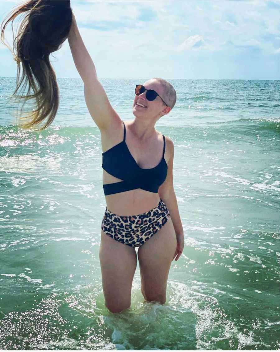 Woman with buzzcut standing in ocean holding out wig