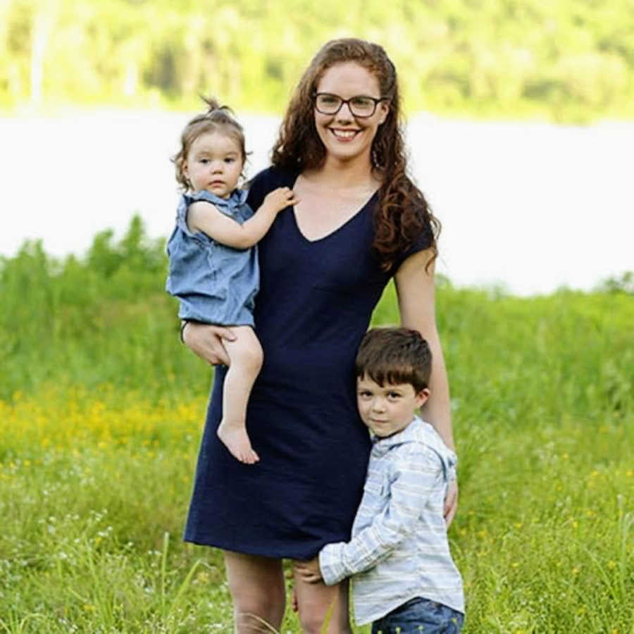 Mother standing in grass by water holding daughter with son grabbing leg