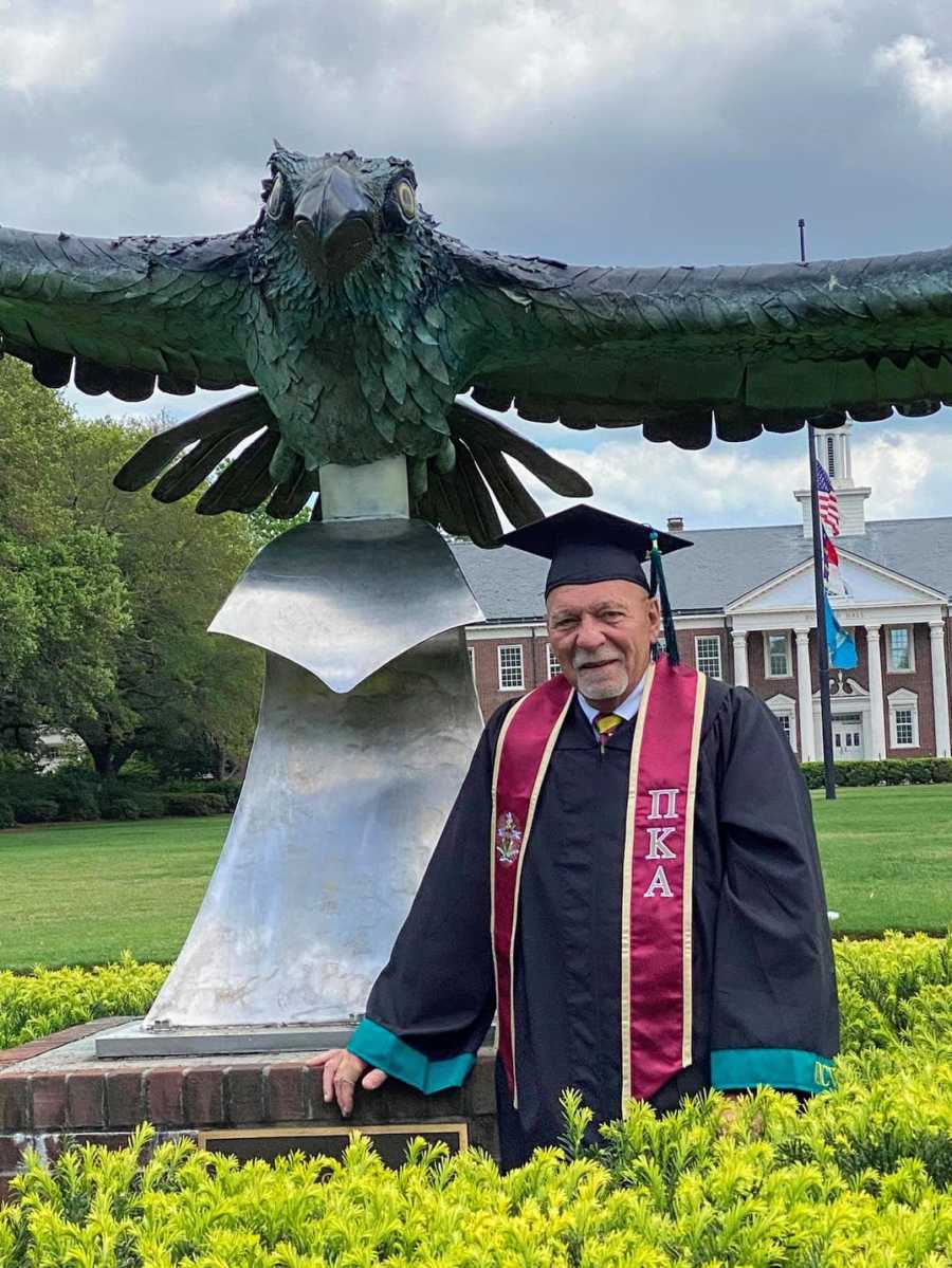 An older college graduate in cap and gown by a bird statue