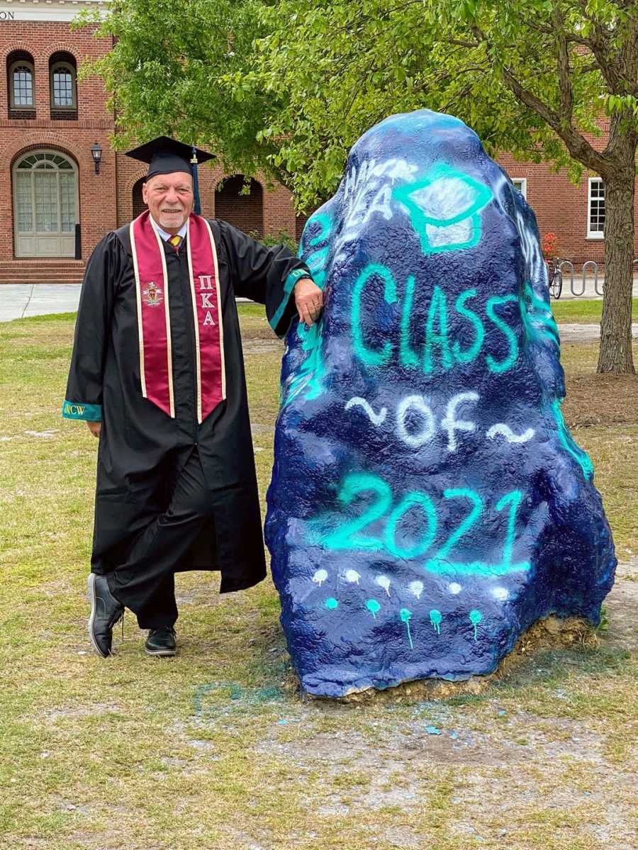 An older college graduate by a rock labeled "Class of 2021"