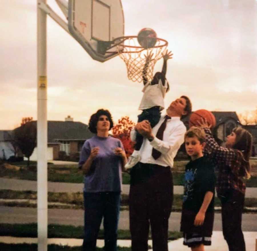 An adoptive father holds up his daughter to shoot a basketball