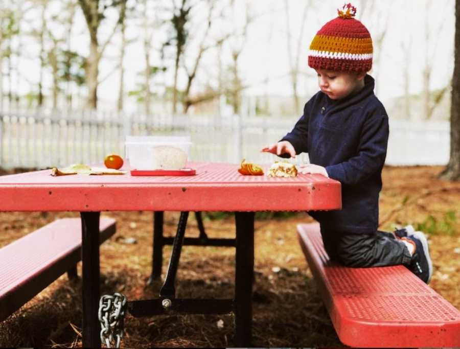 A two-year-old boy sits at a picnic table wearing a hat