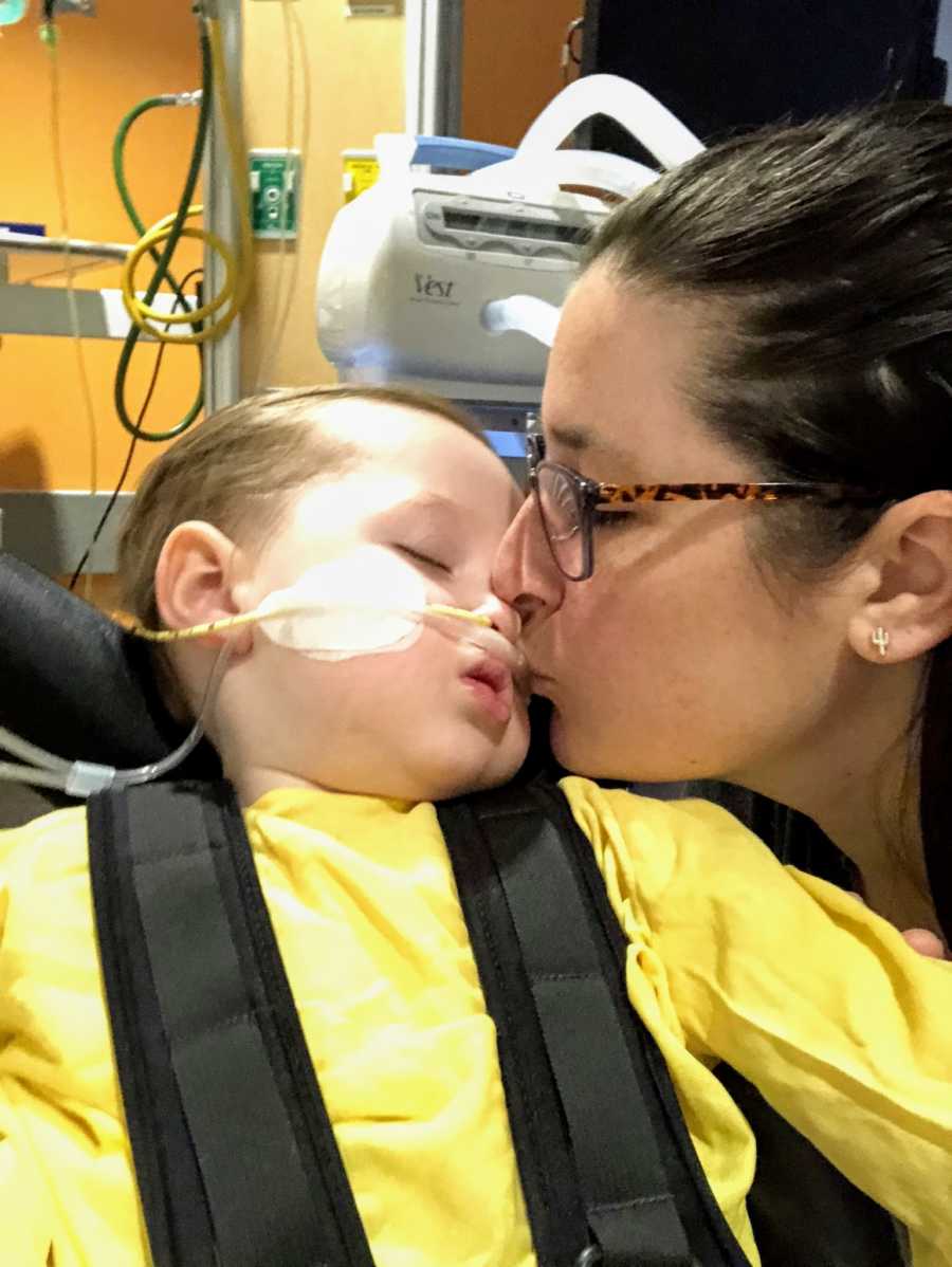 A boy wearing a breathing tube is kissed by his mother