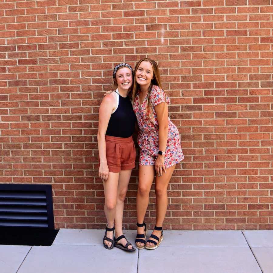 Two friends standing in front of brick wall with arms around each other and smiling