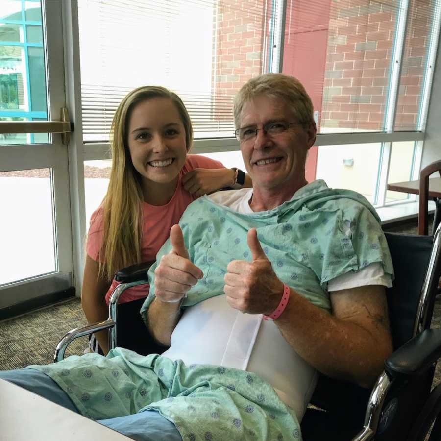 Father wearing hospital gown in wheelchair with daughter doing thumbs up
