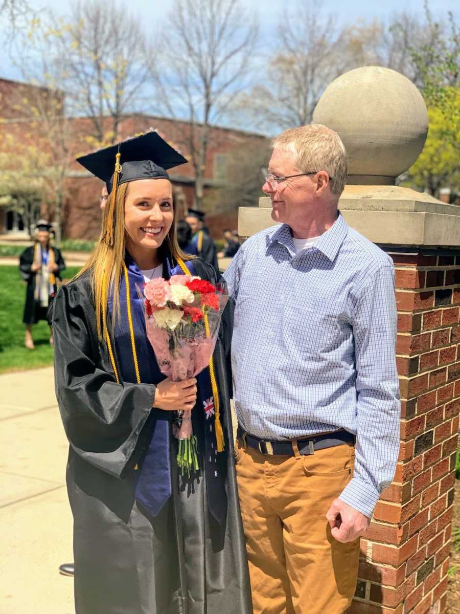 Daughter wearing graduation regalia standing with father and smiling