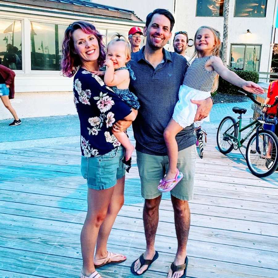 Married couple pose for a family photo with their two daughters on a boardwalk