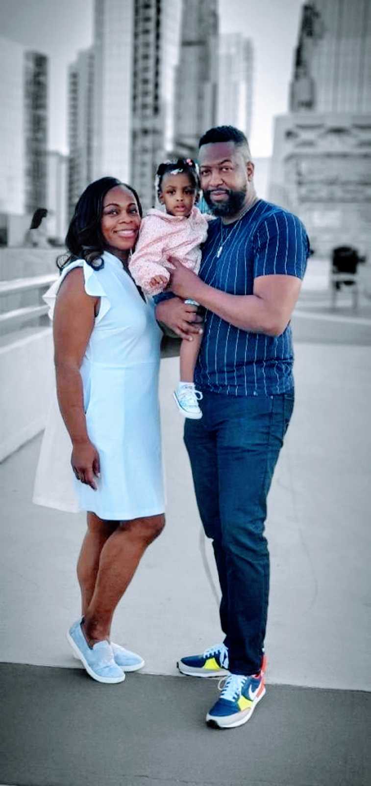 Married couple pose for a photo with their young daughter in front of a city skyline