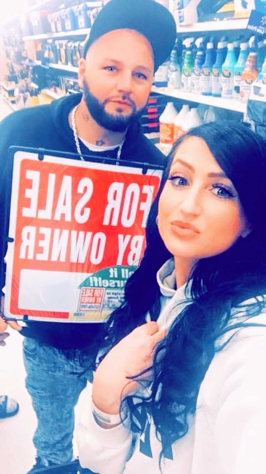 Couple moving in together take a selfie with a "for sale by owner" sign
