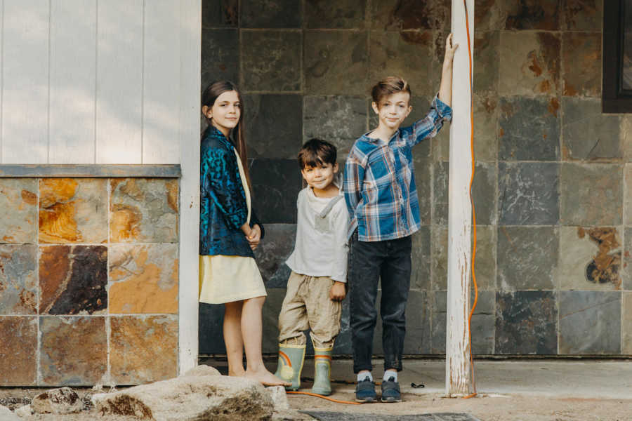 Three siblings standing on porch by stone wall smiling