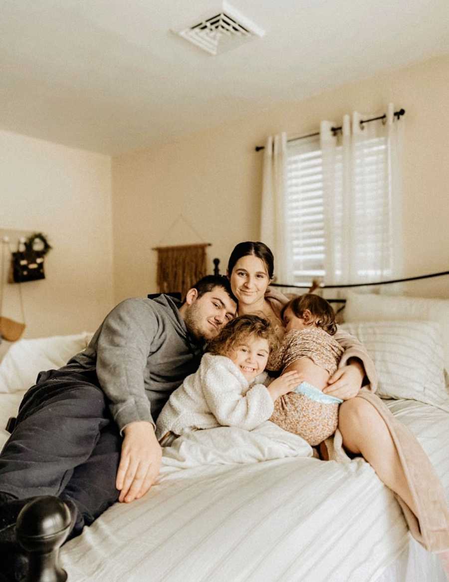 Family of four take a photo together, all piled up in bed together and cuddling