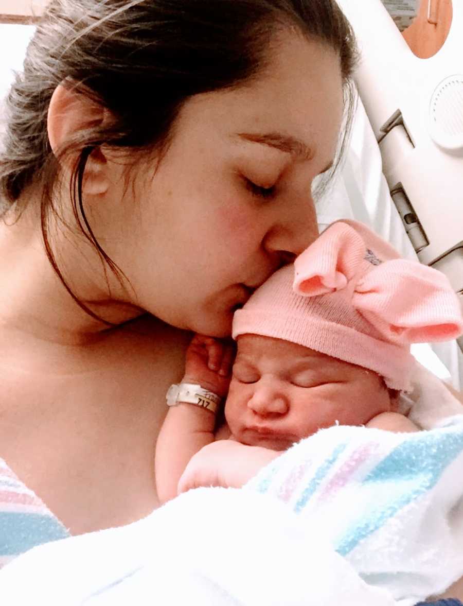 Teen mom takes a photo kissing her newborn daughter on the forehead while laying in a hospital bed after birth