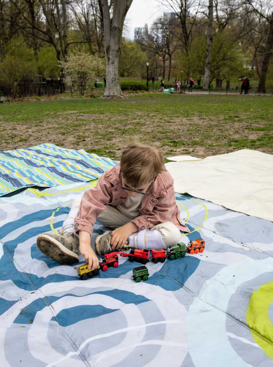 Young boy with glasses sits on a blanket in a park and plays with toy trains