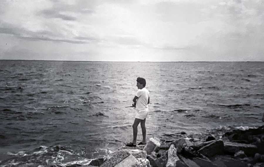 Black and white photo of a man fishing in the ocean while standing on rocks