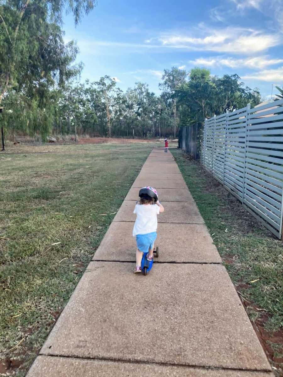 Mom captures her twin three-year-olds riding scooters on a sidewalk with helmets on