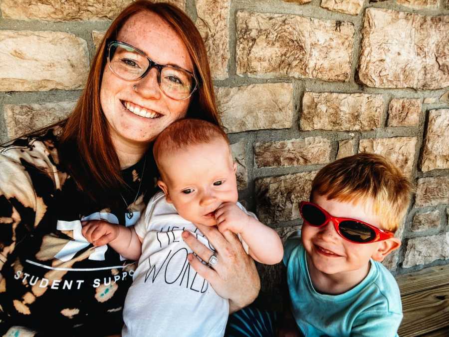 Boy mom holds her newborn son while her toddler sits next to them with red sunglasses on and smiles
