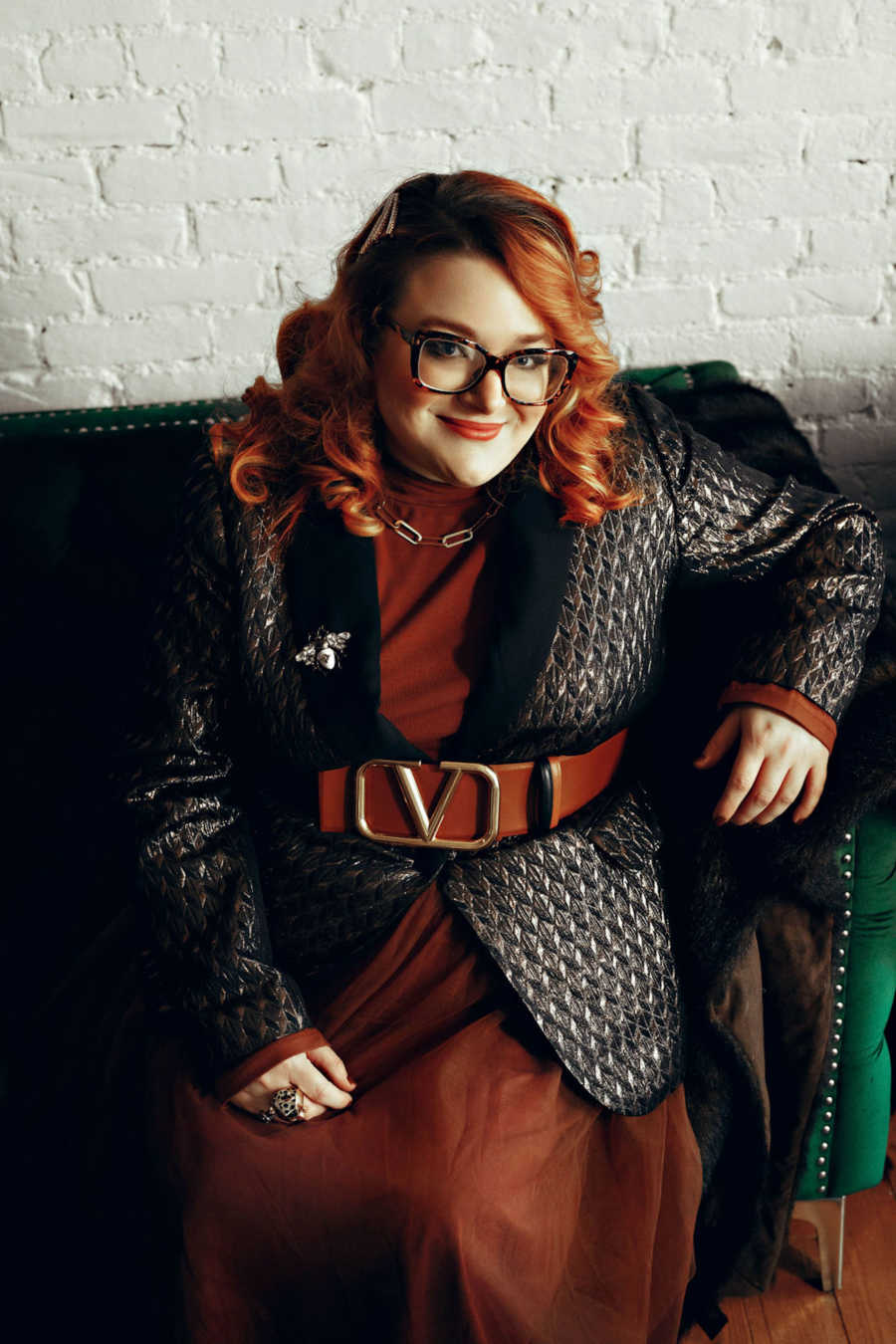 Redhead woman wearing black leather jacket sitting on black couch smiling at camera in front of white brick wall