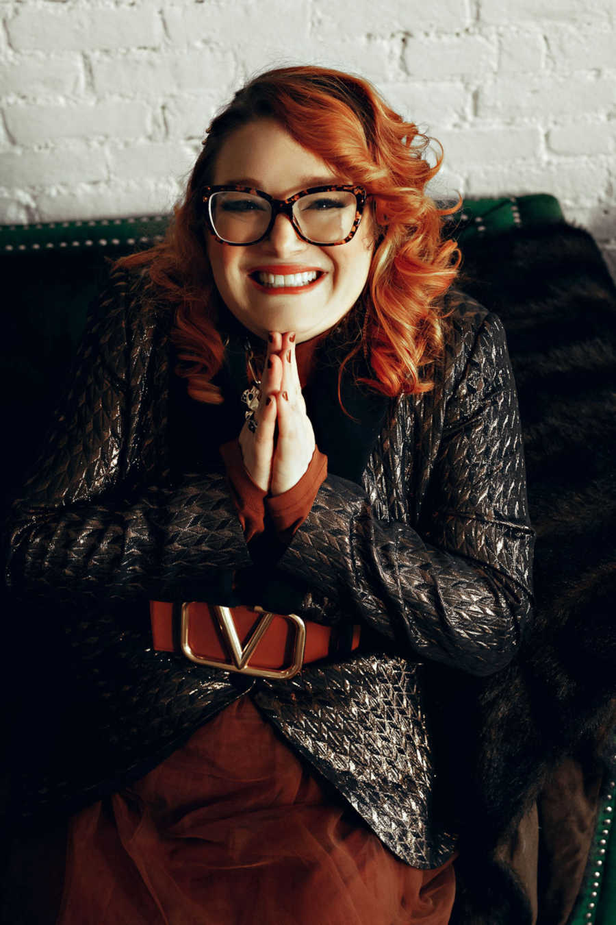 Redhead woman with hands clasped under chin wearing leather jacket sitting on black couch