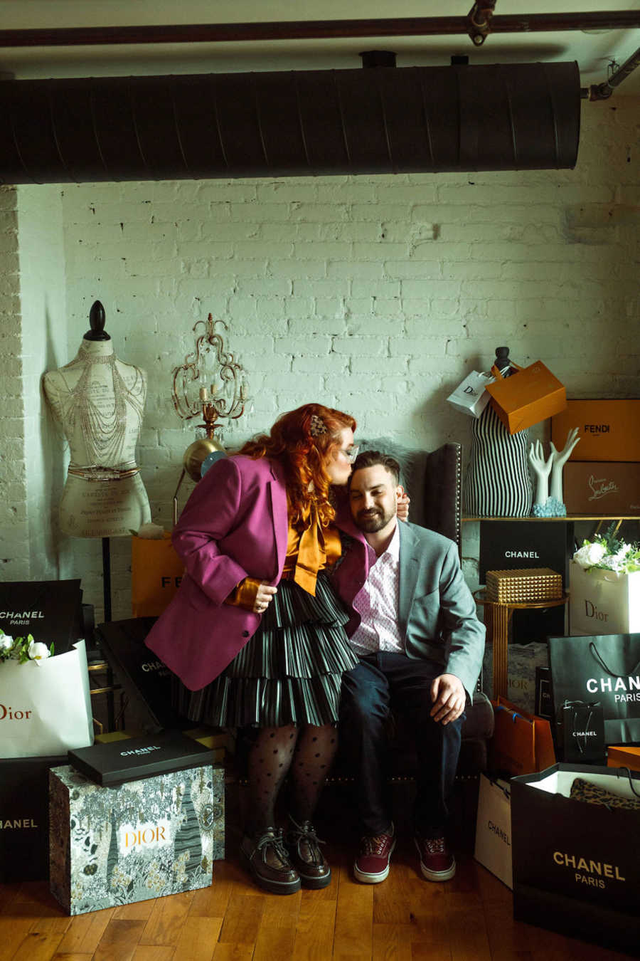 Woman and man sitting in chair surrounded by shopping bags with the woman kissing the man on the head