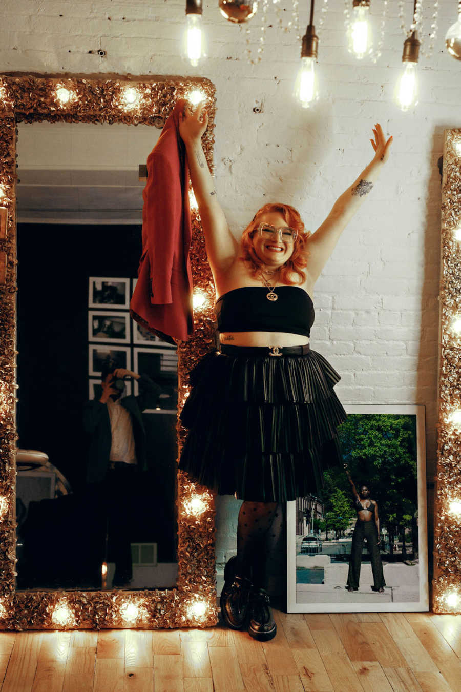 Woman wearing black crop top and black skirt holding arms above head in front of lighted mirror and white brick wall