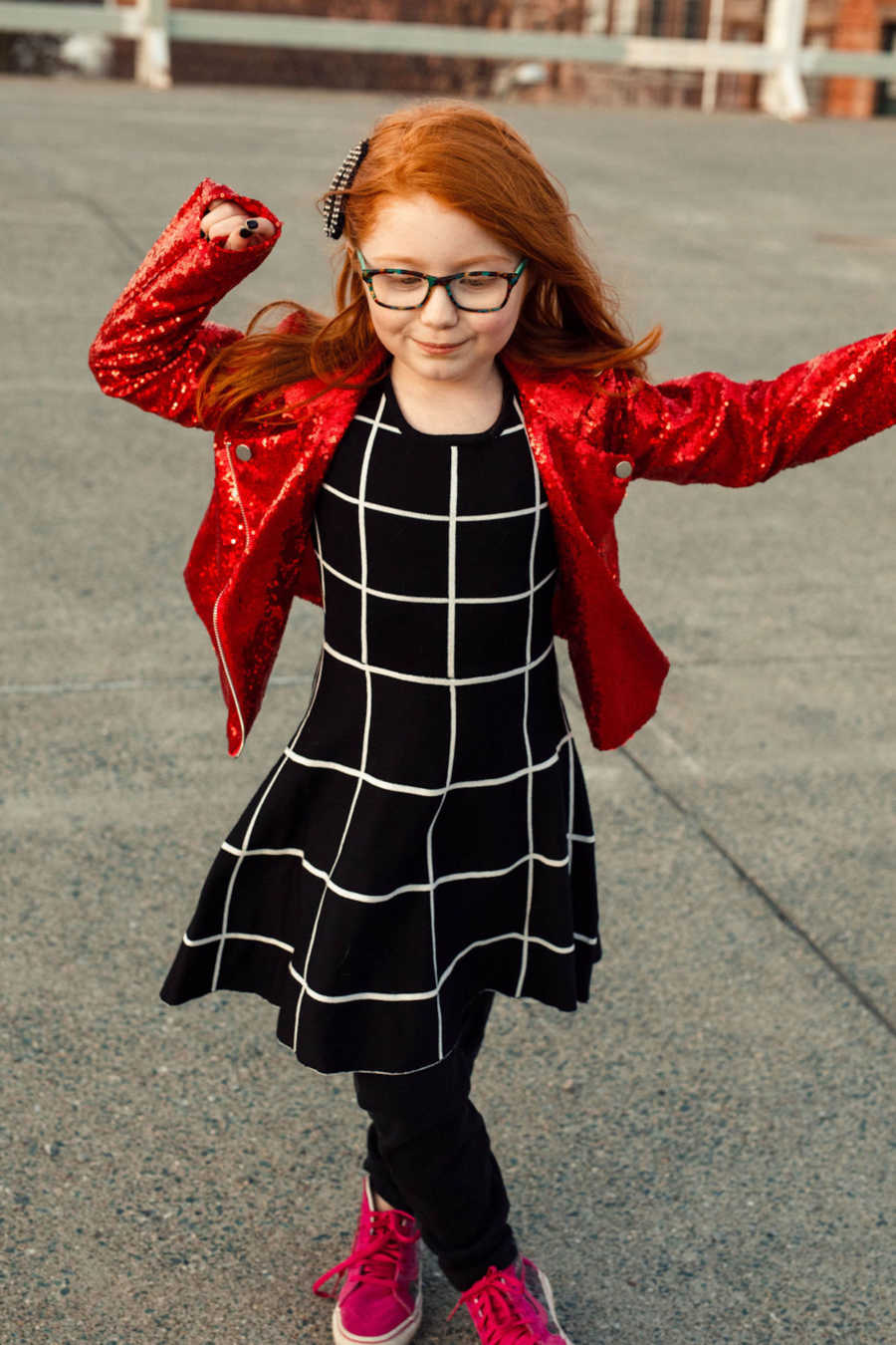 Young redhead girl wearing black grid dress and sparkly red jacket twirling outside