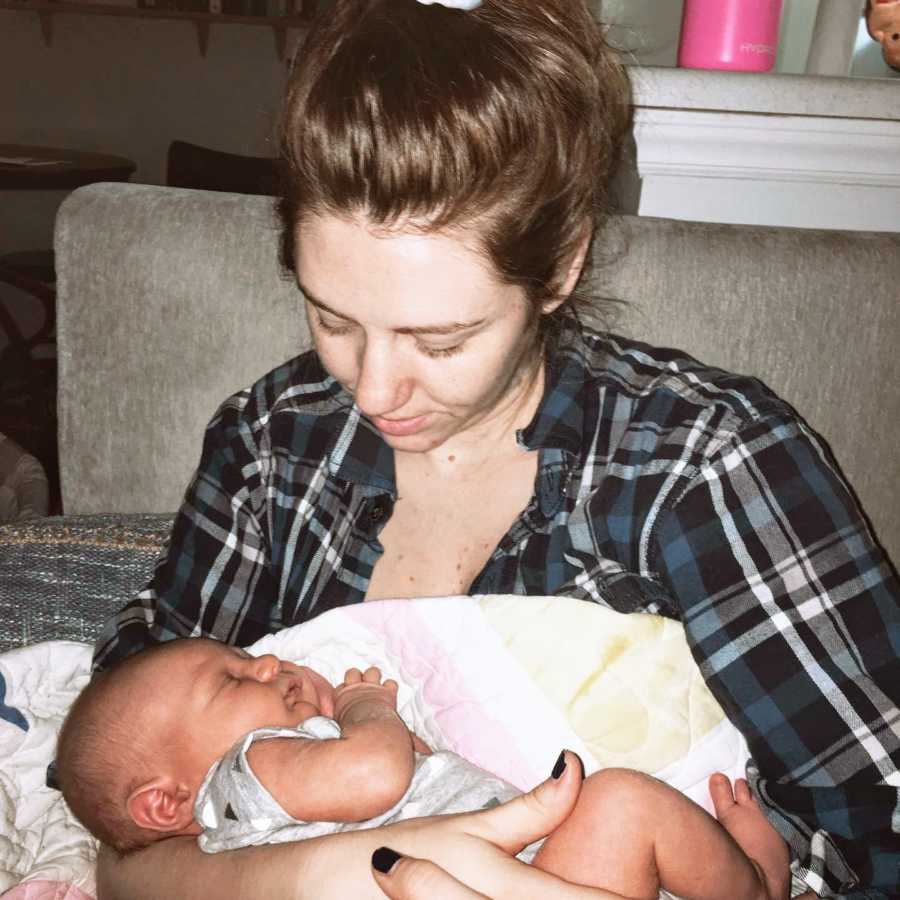 New mom in blue and black plaid shirt smiles down at her sleeping newborn son cradled in her arms