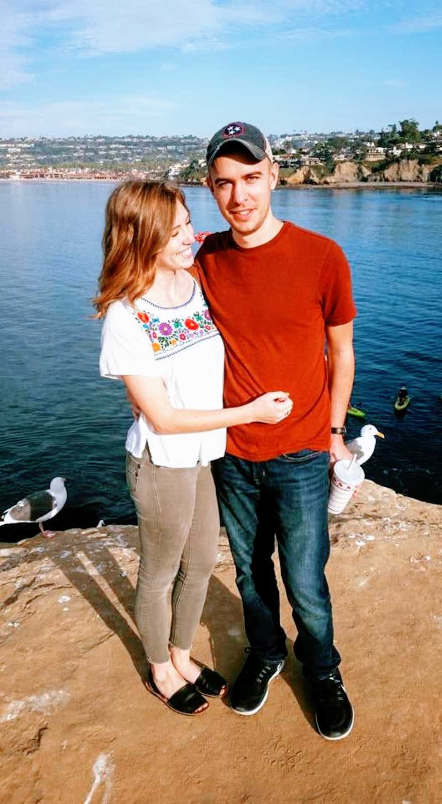 Couple take photo together while standing on a rock near the water with seagulls behind them