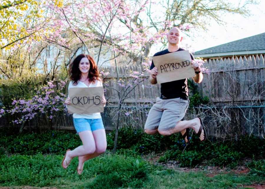 Man and woman jump for joy in their backyard holding signs that say "OKDHS APPROVED"