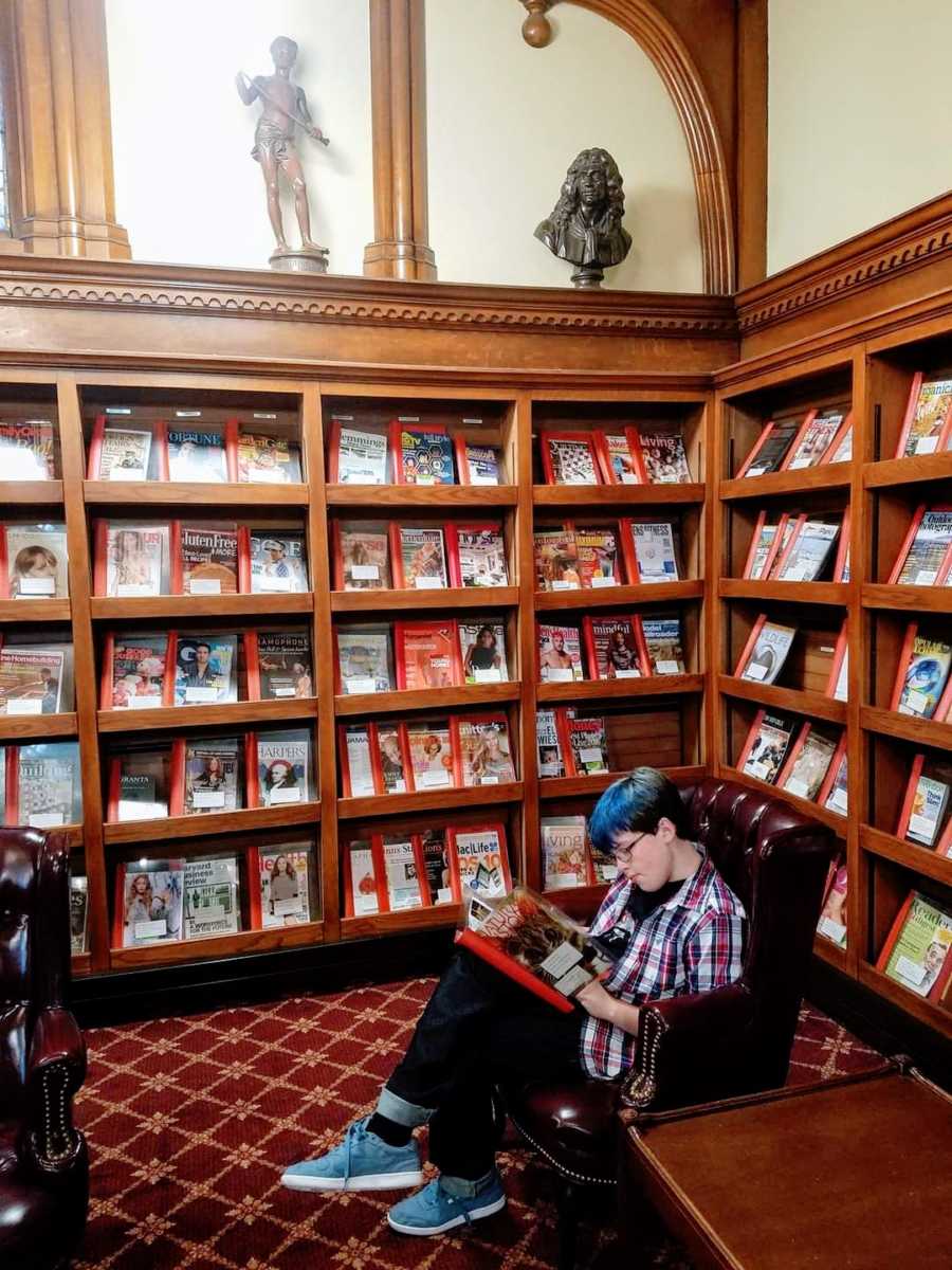 Young boy with blue hair sits in a library, reading a book