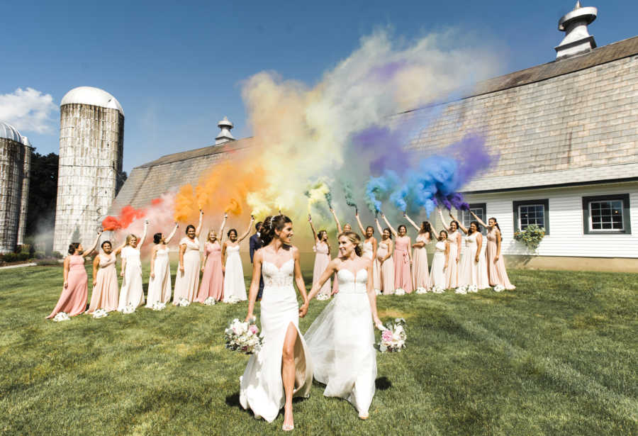 lesbian wedding photo on a hill. bridesmaids in pink and white dresses, rainbow smoke in the air