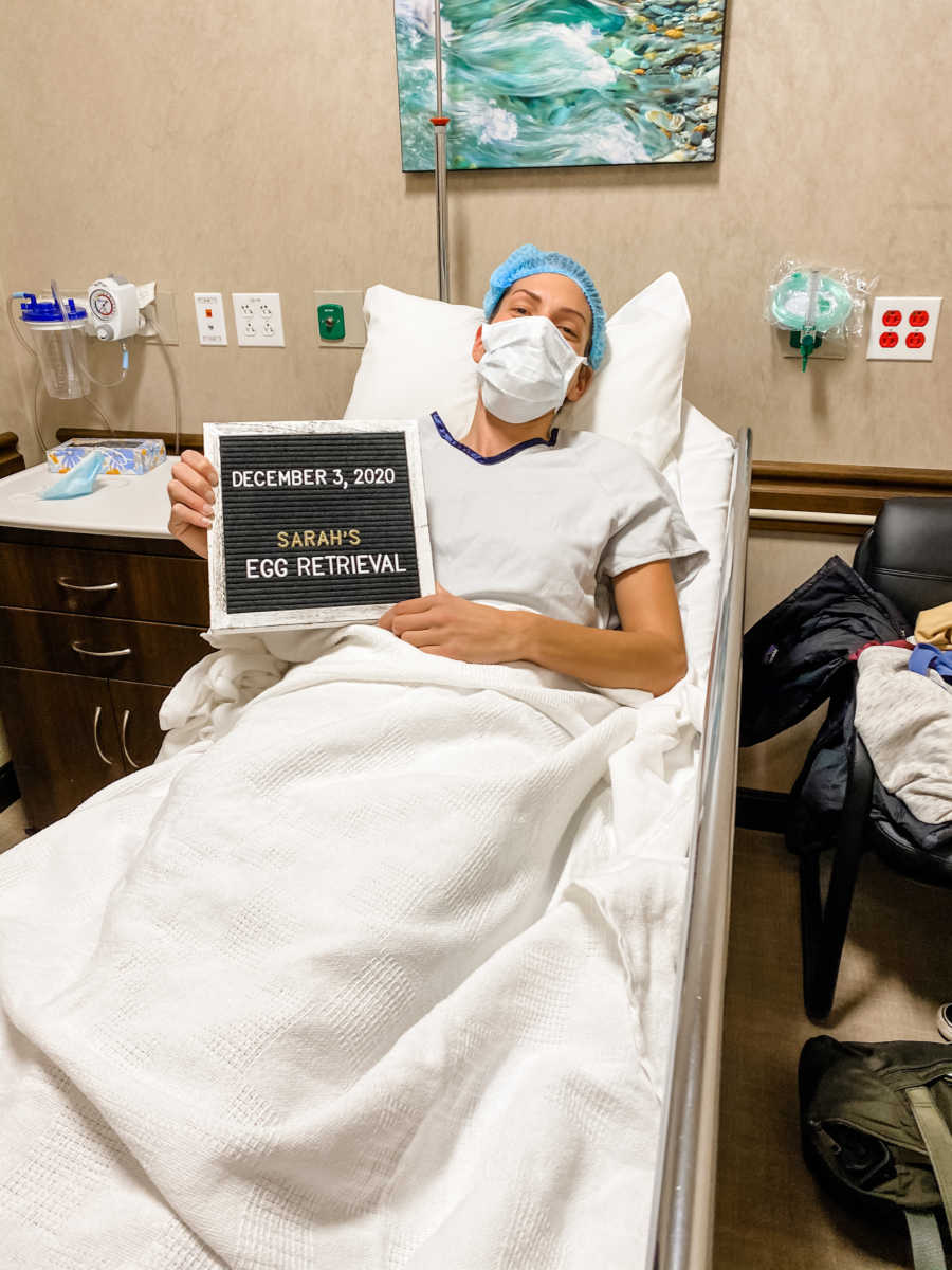 woman in hospital holding sign "Sarah's egg retrieval"