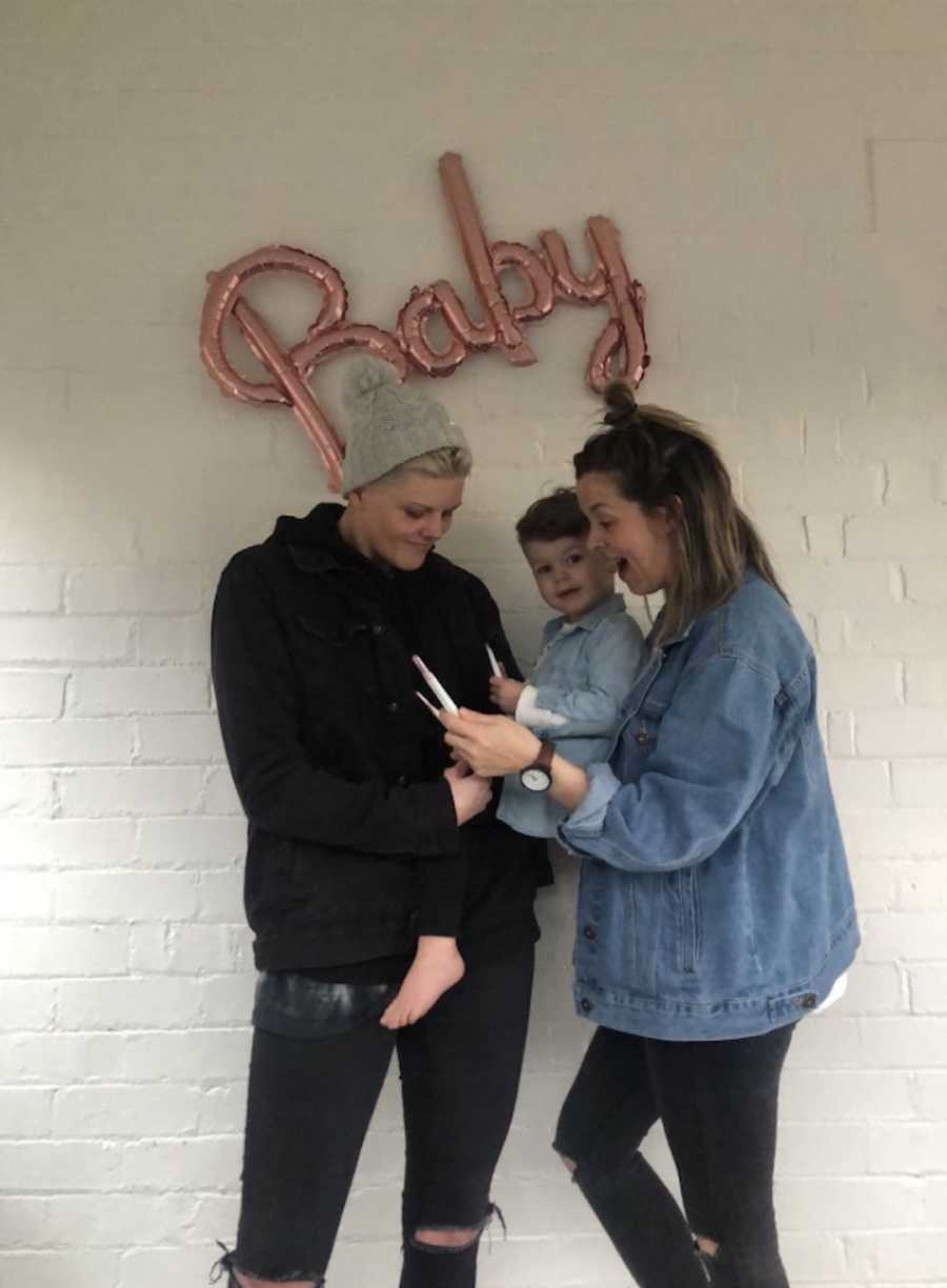 two moms holding baby, "baby" balloon in the background