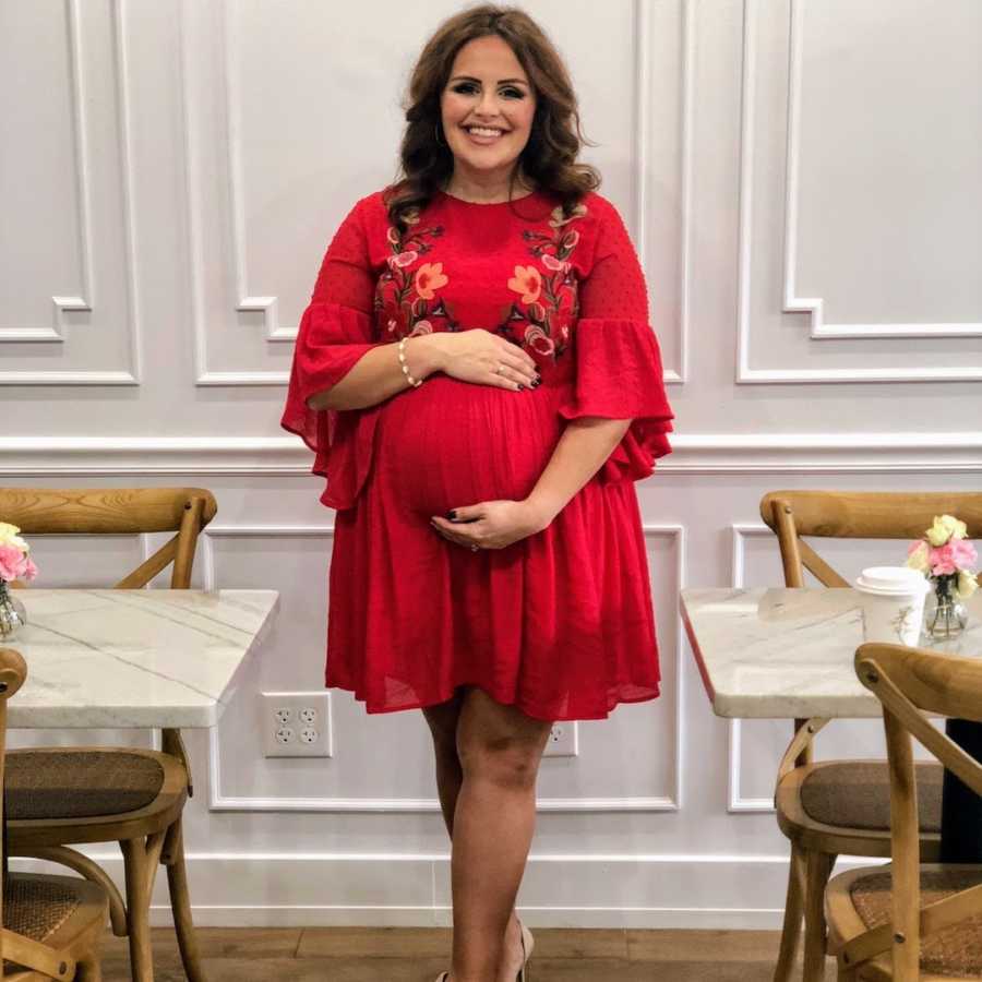 Pregnant woman wearing red dress standing holding belly and smiling