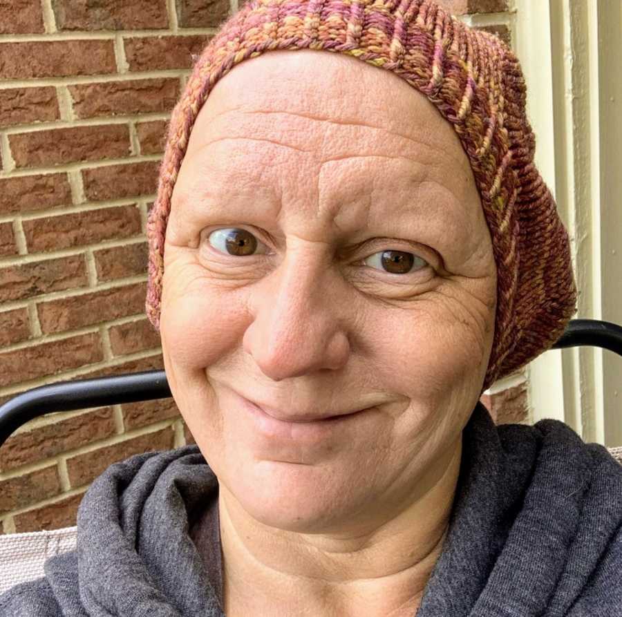 Bald woman wearing beanie taking smiling selfie in front of brick wall