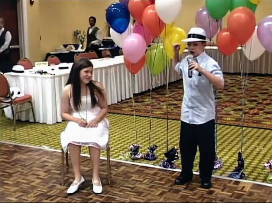 Girl sitting in a chair at birthday party and young boy standing next to her holding a microphone