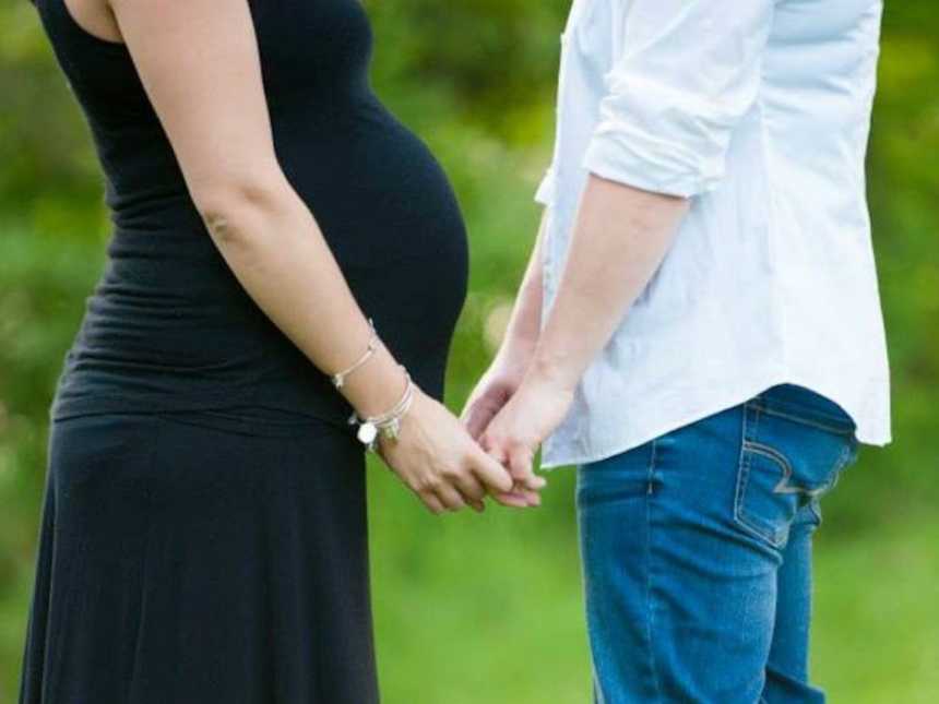 Married wives holding hands, one pregnant and wearing a black dress and the other in jeans and a white shirt